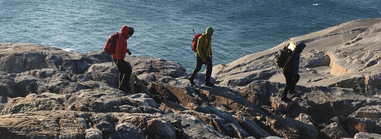 Three hikers with backpacks walk on a rocky shore along water's edge.